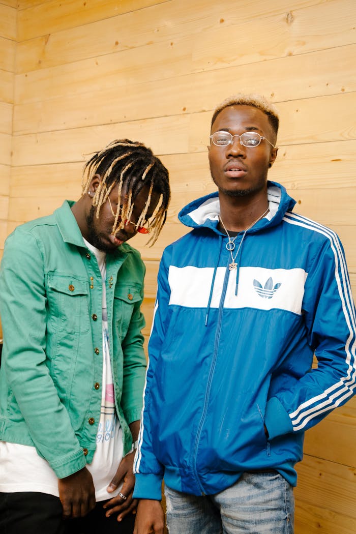 Two stylish young men posing in casual streetwear against a wooden wall.